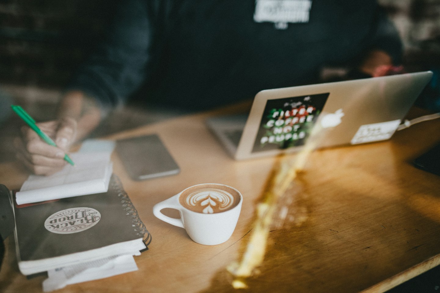 Person schreibt in Notizbuch an Holztisch mit Laptop und Kaffee.